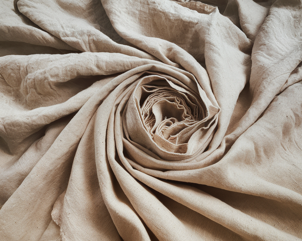 A photo of a large roll of natural linen fabric with a light beige hue. The fabric has a slightly rough texture