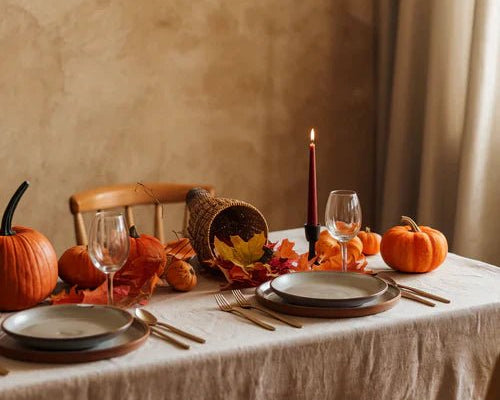 A photograph of a dining table with a linen tablecloth in a fall setting. The table is set with plates, utensils, and a candle. There are pumpkins, leaves, and a cornucopia on the table.