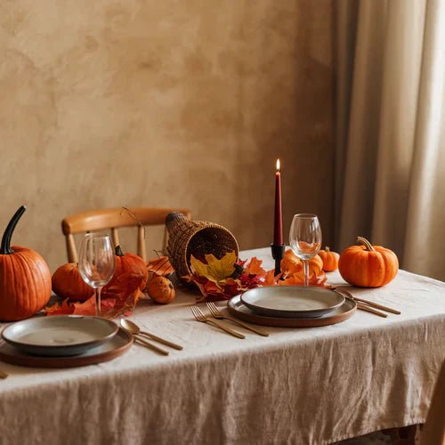A photograph of a dining table with a linen tablecloth in a fall setting. The table is set with plates, utensils, and a candle. There are pumpkins, leaves, and a cornucopia on the table.