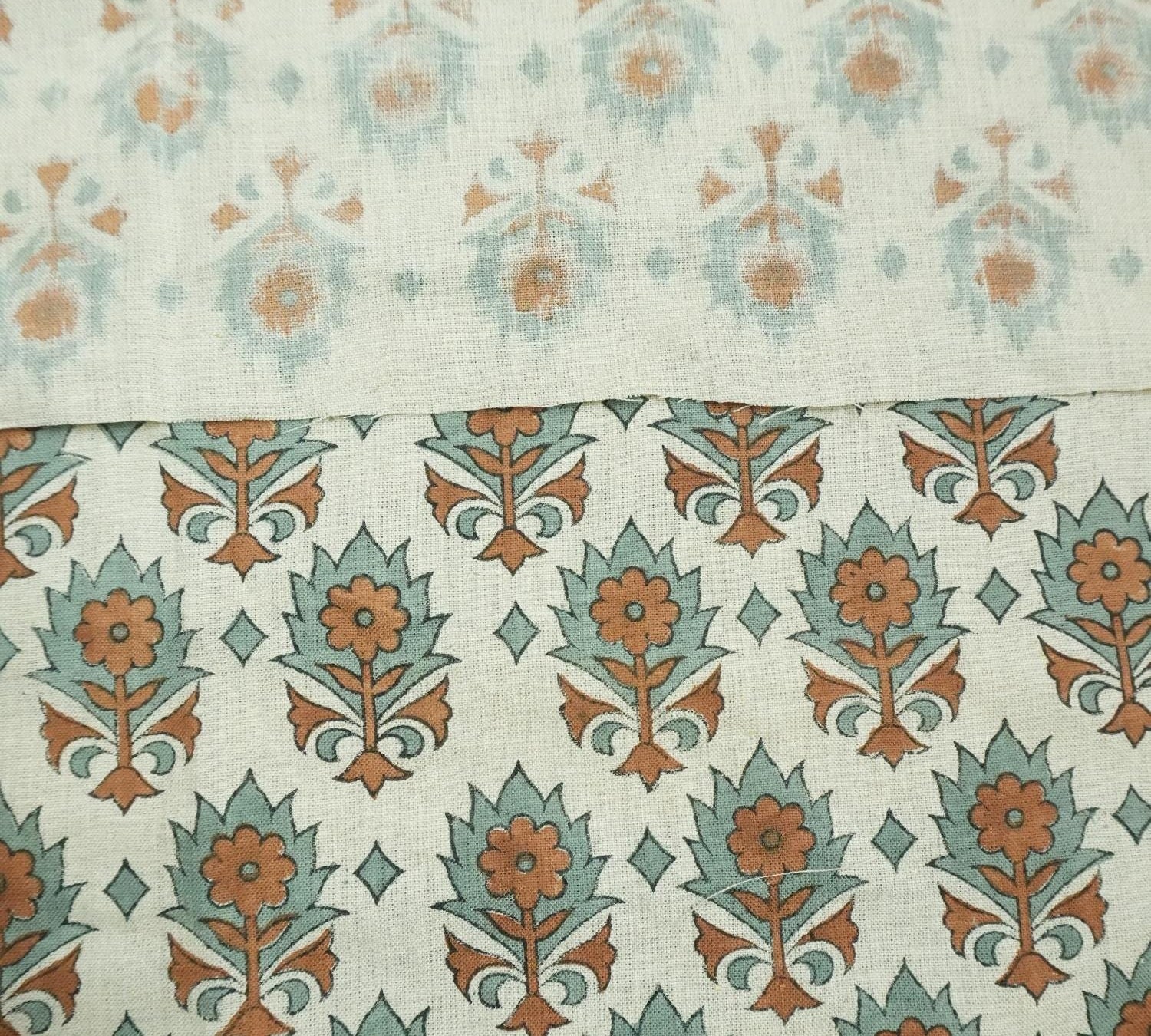 May include: Close-up view of two fabric swatches showcasing a subtle, repeating floral pattern. The top swatch displays a lighter, more faded pattern in muted teal and beige, while the bottom swatch features a bolder, more defined pattern in teal and burnt orange. Both fabrics share a similar base color of off-white or beige linen, creating a cohesive design. The patterns are reminiscent of traditional block-printed textiles, suggesting a handcrafted or vintage aesthetic.