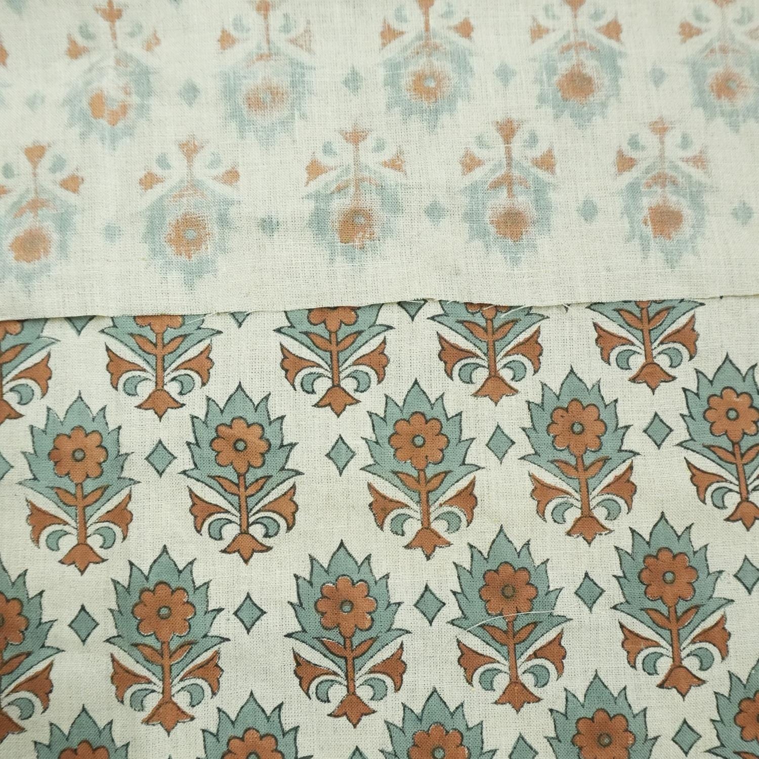 May include: Close-up view of two fabric swatches showcasing a subtle, repeating floral pattern. The top swatch displays a lighter, more faded pattern in muted teal and beige, while the bottom swatch features a bolder, more defined pattern in teal and burnt orange. Both fabrics share a similar base color of off-white or beige linen, creating a cohesive design. The patterns are reminiscent of traditional block-printed textiles, suggesting a handcrafted or vintage aesthetic.